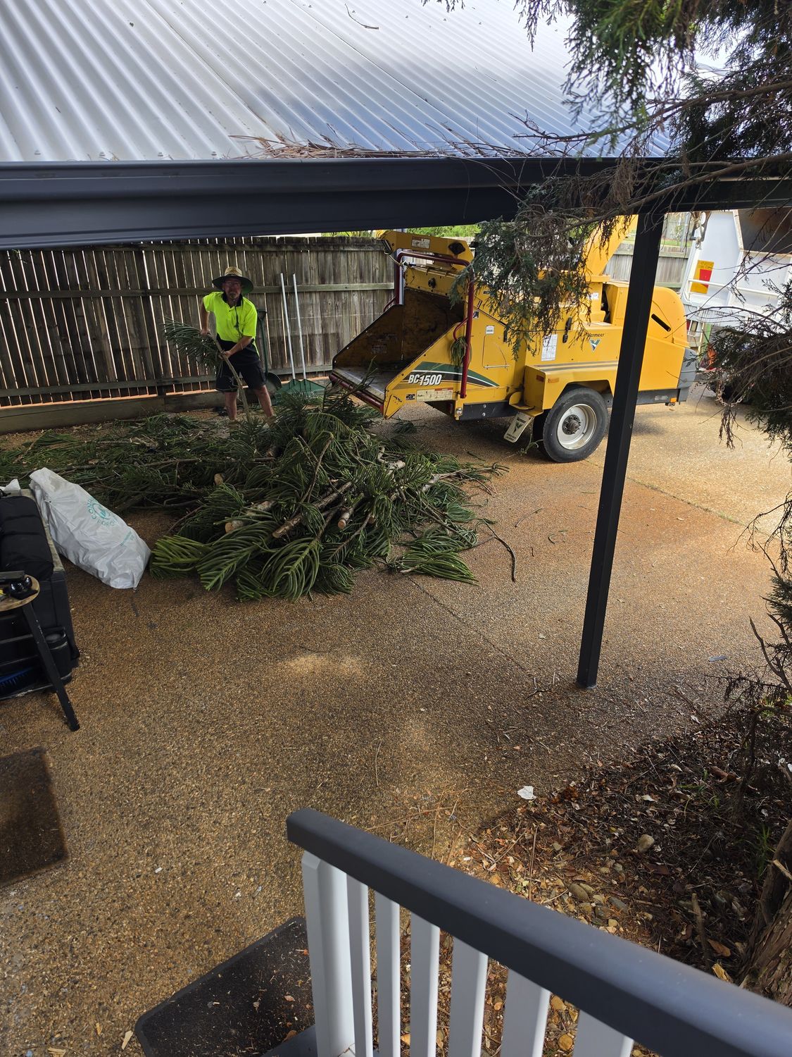 Crew member feeding branches into a yellow wood chipper beside a carport